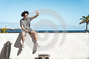 Young Latin man having fun taking selfie with mobile smartphone while listening music with headphones and boombox during summer