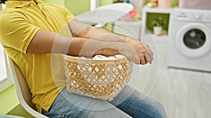 Young latin man holding basket with clothes sitting on chair at laundry room