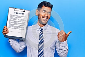 Young latin man holding clipboard with contract document pointing thumb up to the side smiling happy with open mouth