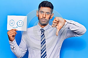 Young latin man holding sad to happy emotion paper with angry face, negative sign showing dislike with thumbs down, rejection