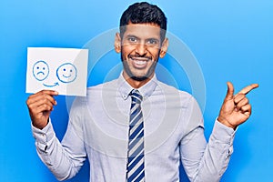 Young latin man holding sad to happy emotion paper smiling happy pointing with hand and finger to the side