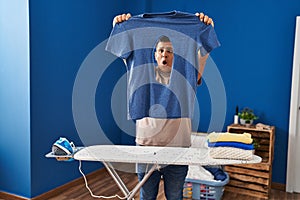 Young latin man holding t shirt with burned hole with surprise expression at laundry room
