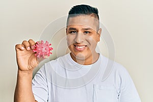Young latin man holding virus toy looking positive and happy standing and smiling with a confident smile showing teeth