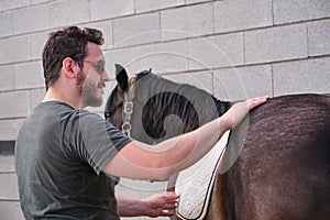 Young latin man putting on the saddle blanket to his horse and stroking it.