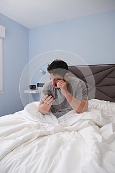 Young latin man rubbing his eyes in bed after sleeping and checking his smartphone