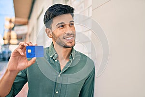 Young latin man smiling happy holding credit card leaning on the wall at the city