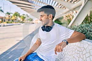 Young latin man smiling happy listening to music using headphones sitting on the bench at the park