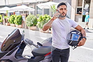 Young latin man talking on smartphone standing by motorbike at street