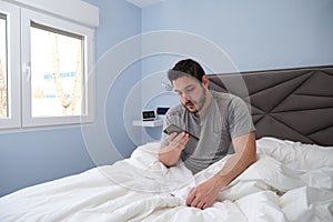 Young latin man tired in bed after sleeping and checking his smartphone
