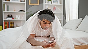 Young latin man using smartphone covering with blanket surprised at bedroom