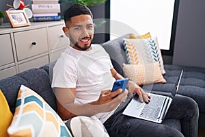 Young latin man using smartphone and laptop sitting on sofa at home
