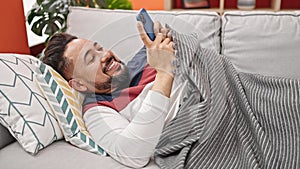 Young latin man using smartphone lying on sofa at home