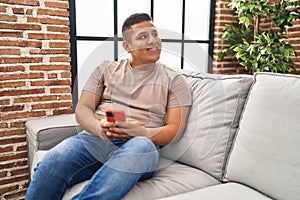 Young latin man using smartphone sitting on sofa at home