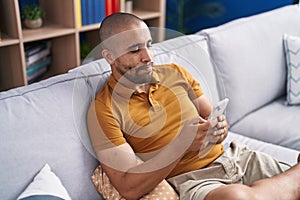 Young latin man using smartphone sitting on sofa at home