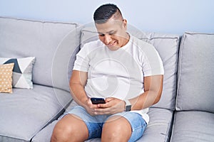 Young latin man using smartphone sitting on sofa at home