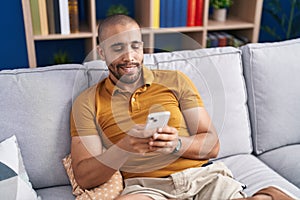 Young latin man using smartphone sitting on sofa at home