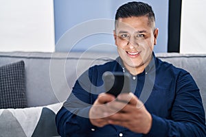 Young latin man using smartphone sitting on sofa at home