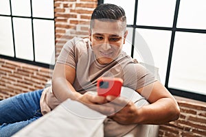 Young latin man using smartphone sitting on sofa at home