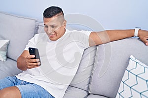 Young latin man using smartphone sitting on sofa at home