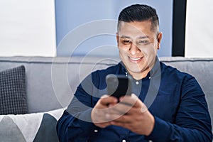 Young latin man using smartphone sitting on sofa at home