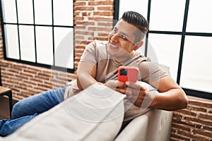 Young latin man using smartphone sitting on sofa at home