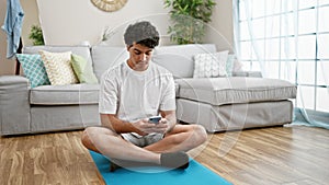 Young latin man using smartphone sitting on yoga mat at home