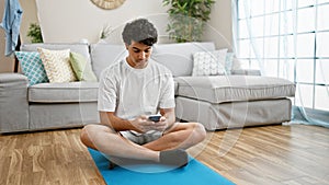 Young latin man using smartphone sitting on yoga mat at home