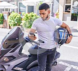 Young latin man using smartphone standing by motorbike at street