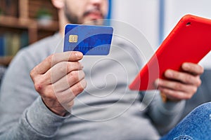 Young latin man using touchpad and credit card sitting on sofa at home