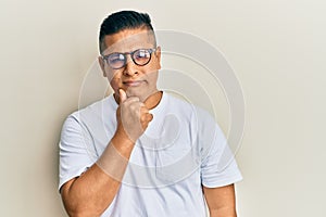 Young latin man wearing casual clothes and glasses thinking concentrated about doubt with finger on chin and looking up wondering