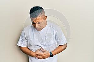 Young latin man wearing casual white t shirt with hand on stomach because indigestion, painful illness feeling unwell