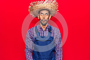 Young latin man wearing farmer hat and apron making fish face with lips, crazy and comical gesture