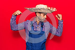 Young latin man wearing farmer hat and apron showing arms muscles smiling proud