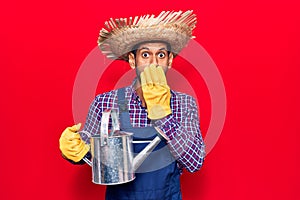 Young latin man wearing farmer hat and gloves holding watering can covering mouth with hand, shocked and afraid for mistake