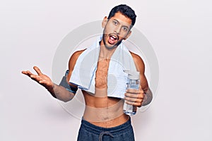 Young latin man wearing sportswear and towel holding bottle of water celebrating achievement with happy smile and winner