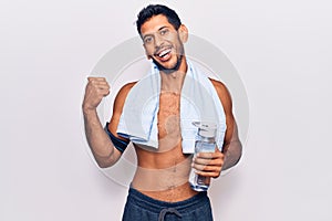 Young latin man wearing sportswear and towel holding bottle of water pointing thumb up to the side smiling happy with open mouth