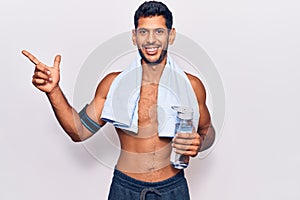 Young latin man wearing sportswear and towel holding bottle of water smiling happy pointing with hand and finger to the side