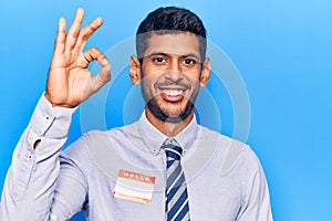 Young latin man wearing sticker with hello my name is message doing ok sign with fingers, smiling friendly gesturing excellent