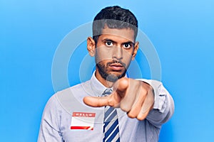 Young latin man wearing sticker with hello my name is message pointing with finger to the camera and to you, confident gesture