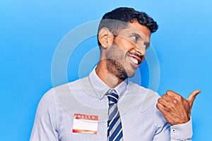 Young latin man wearing sticker with hello my name is message pointing thumb up to the side smiling happy with open mouth
