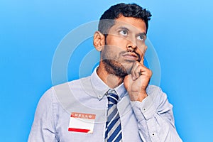 Young latin man wearing sticker with hello my name is message serious face thinking about question with hand on chin, thoughtful