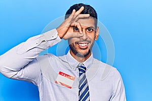 Young latin man wearing sticker with hello my name is message smiling happy doing ok sign with hand on eye looking through fingers