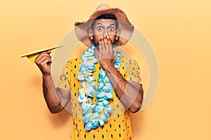 Young latin man wearing summer hat and hawaiian lei holding paper airplane covering mouth with hand, shocked and afraid for