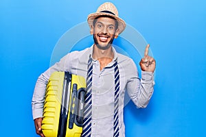 Young latin man wearing summer hat holding cabin bag surprised with an idea or question pointing finger with happy face, number