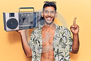 Young latin man wearing summer shirt holding boombox surprised with an idea or question pointing finger with happy face, number