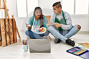 Young latin painter couple having online paint class using laptop sitting on the floor at art studio