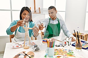 Young latin painter couple smiling happy using manikin sitting on the table at art studio