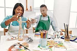 Young latin painter couple smiling happy using manikin sitting on the table at art studio