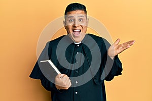 Young latin priest man holding bible celebrating achievement with happy smile and winner expression with raised hand