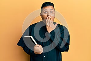 Young latin priest man holding bible covering mouth with hand, shocked and afraid for mistake